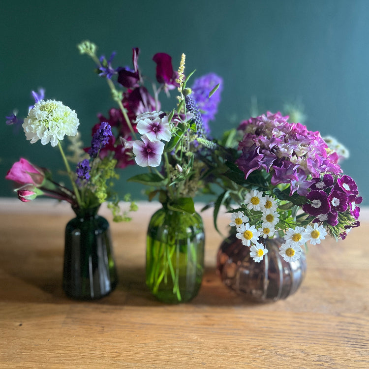 Three small vases with assorted flowers on a wooden surface against a green background