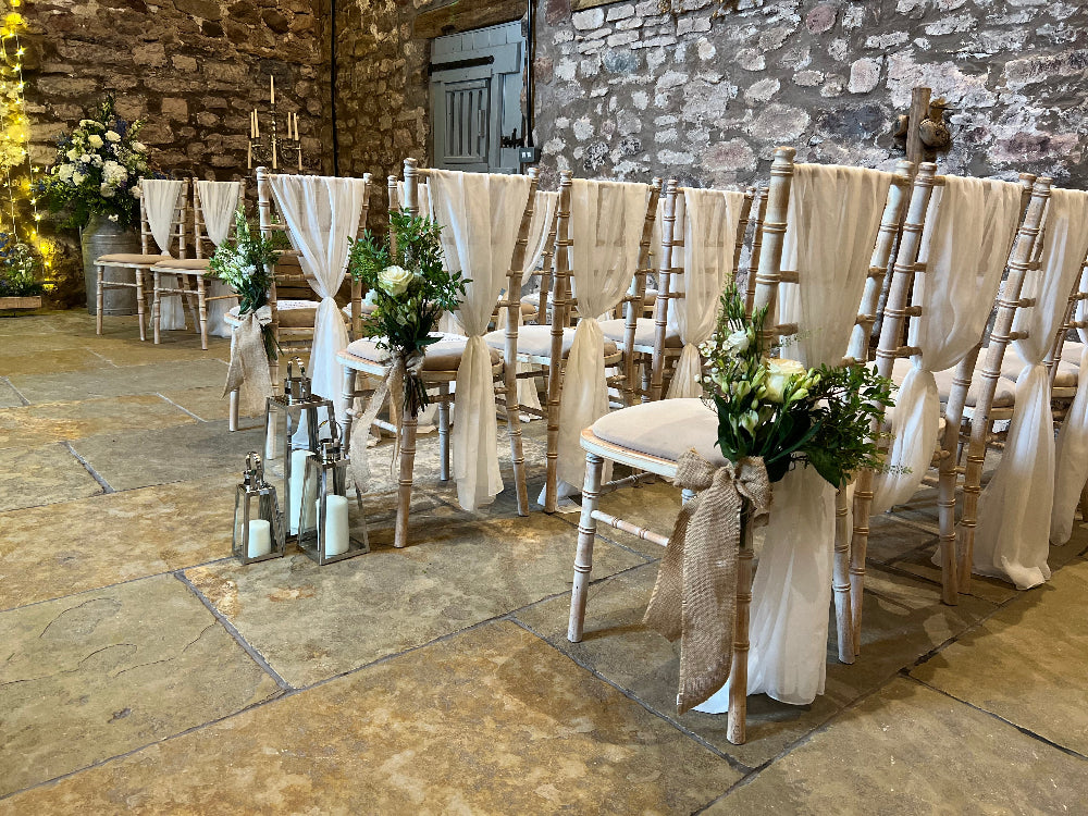 Decorated chairs with white ribbons and floral arrangements in a stone-walled room.