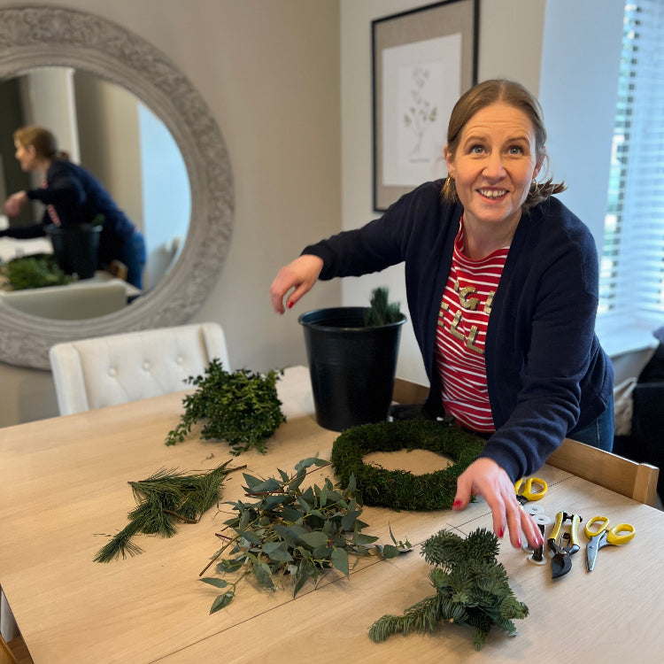 Woman making a wreath at a table with various greenery and tools.