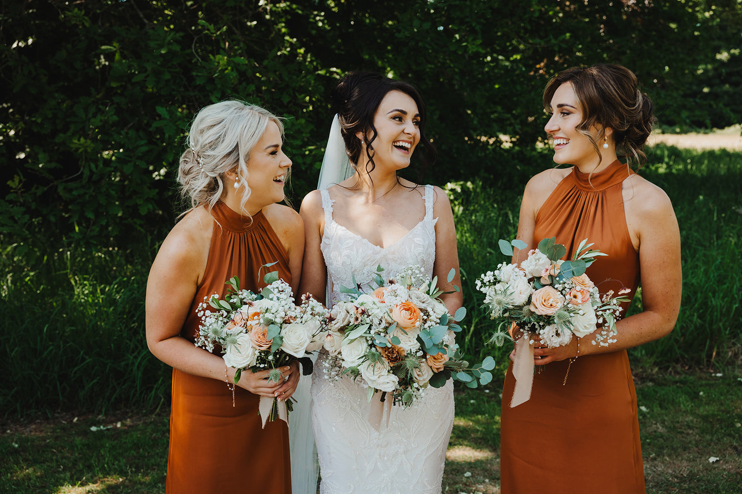 Bride with two bridesmaids holding floral bouquets outdoors.