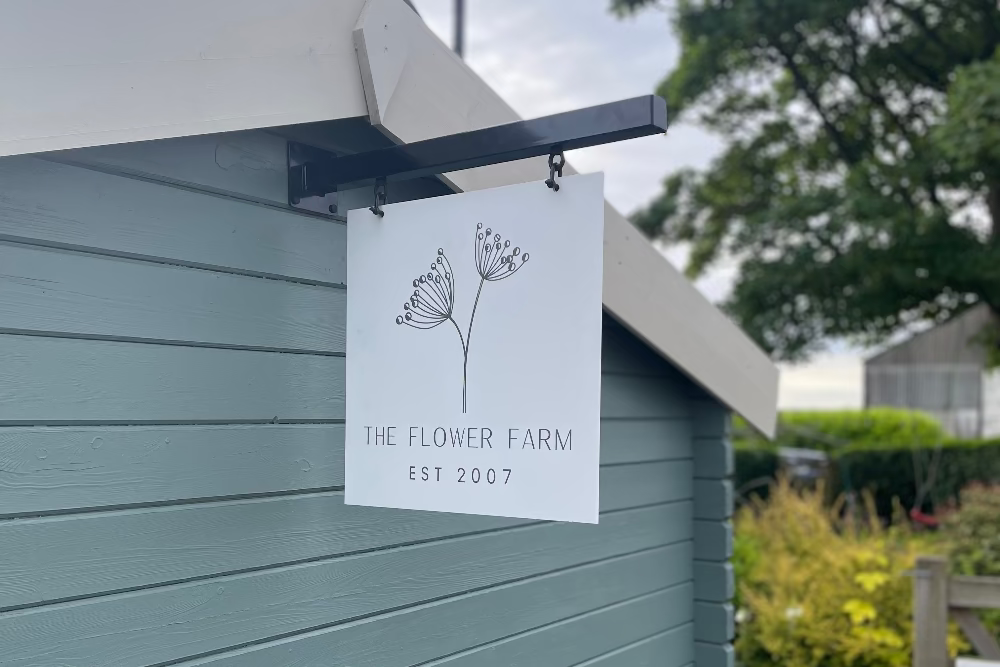 Sign for 'The Flower Farm' on a building exterior with a garden background.
