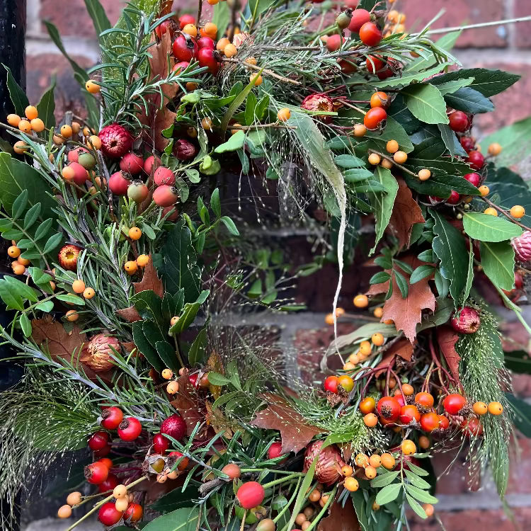 A fresh decorative wreath with greenery, berries, and leaves against a brick wall.