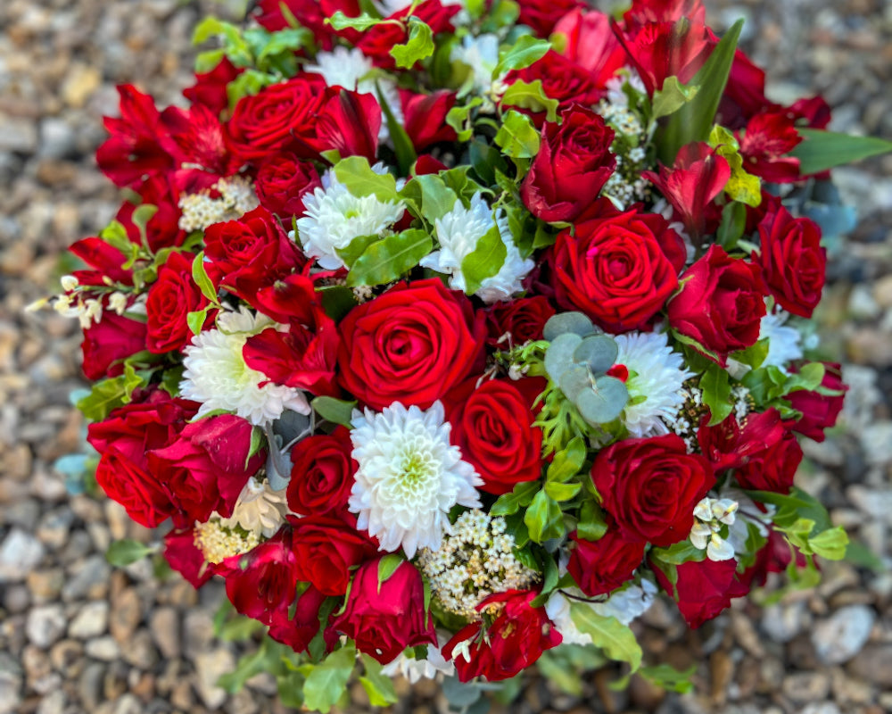 A round funeral tribute created using beautiful red roses, green foliage and white Chrysanthemums