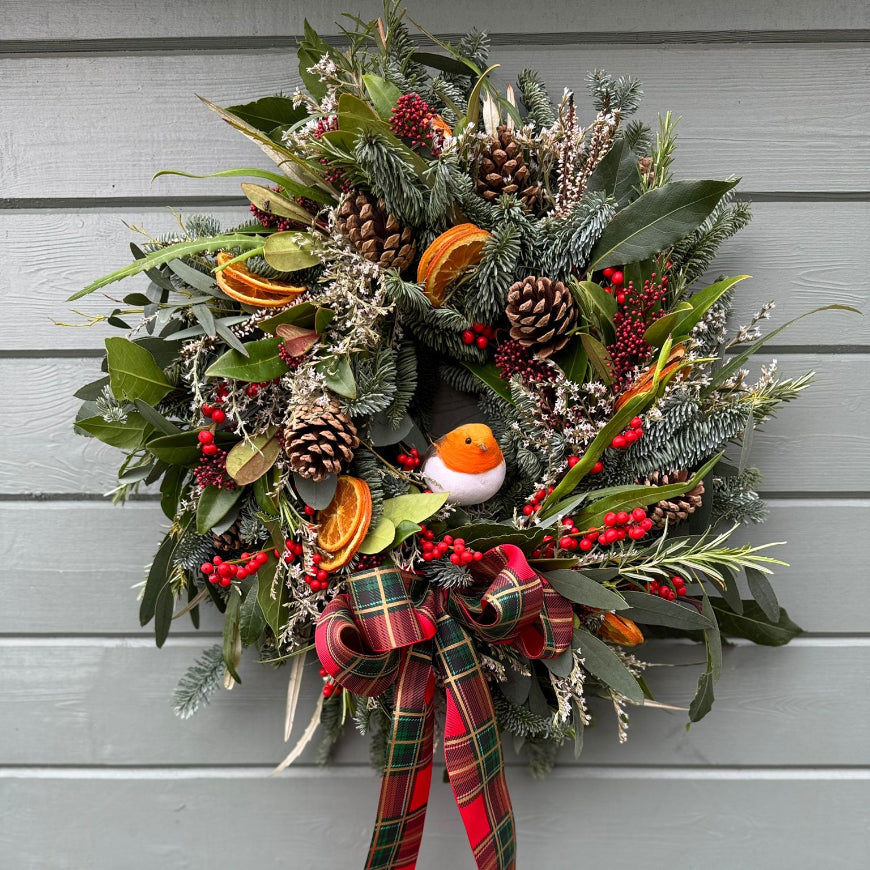 Our nature Inspired Christmas wreath from Flower Farm Floristry, displayed on the outside wall of our log cabin workshop. This wreath features a robin, tartan bow, orange slices, Ilex berries, skimmia, spruce, limonium, fir cones, rosemary, eucalyptus, bay.