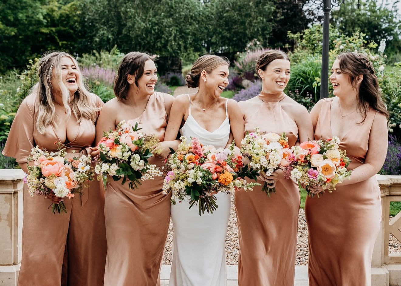 Bride in a white dress with four bridesmaids in brown dresses holding floral bouquets outdoors.